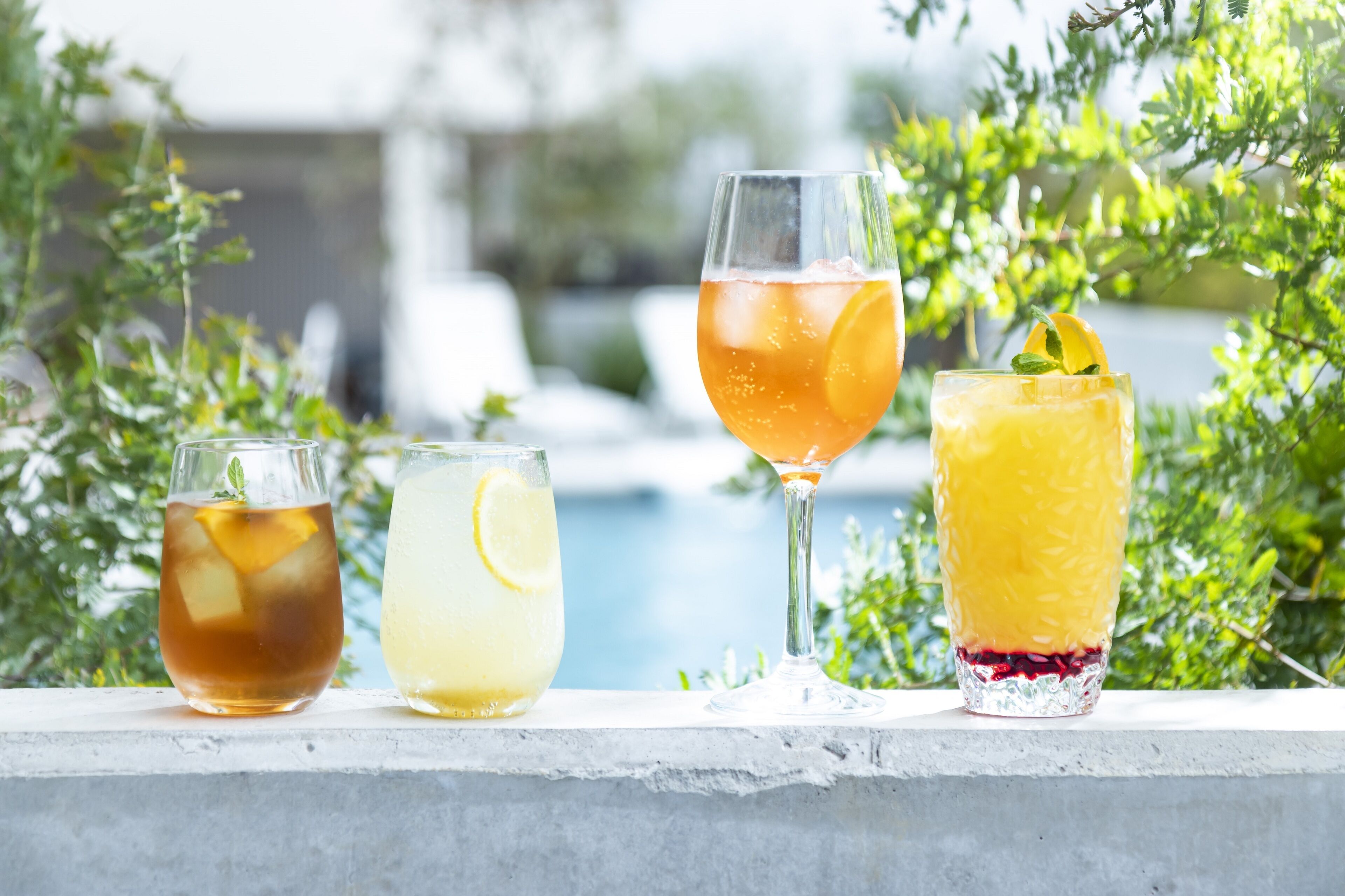 Three glasses of beverages on a ledge with a pool and greenery in the background.
