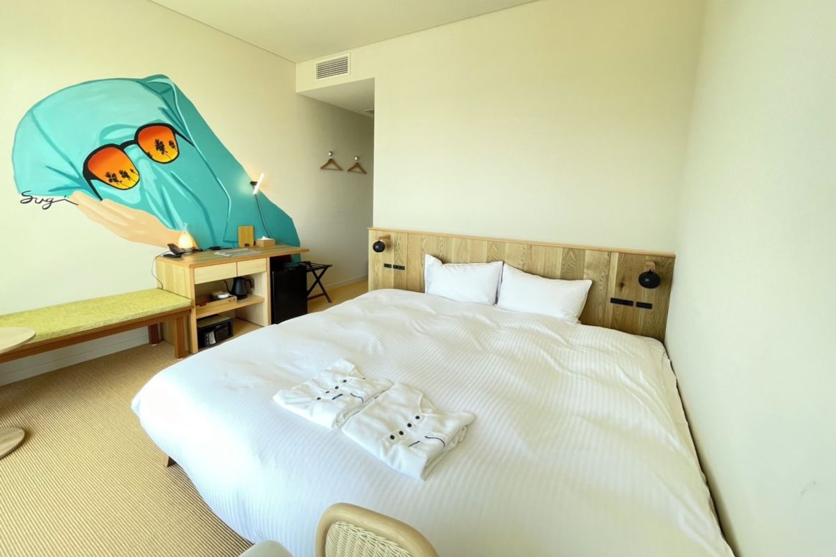 A hotel room with a bed, a desk, a chair, and a wall-mounted shelf.