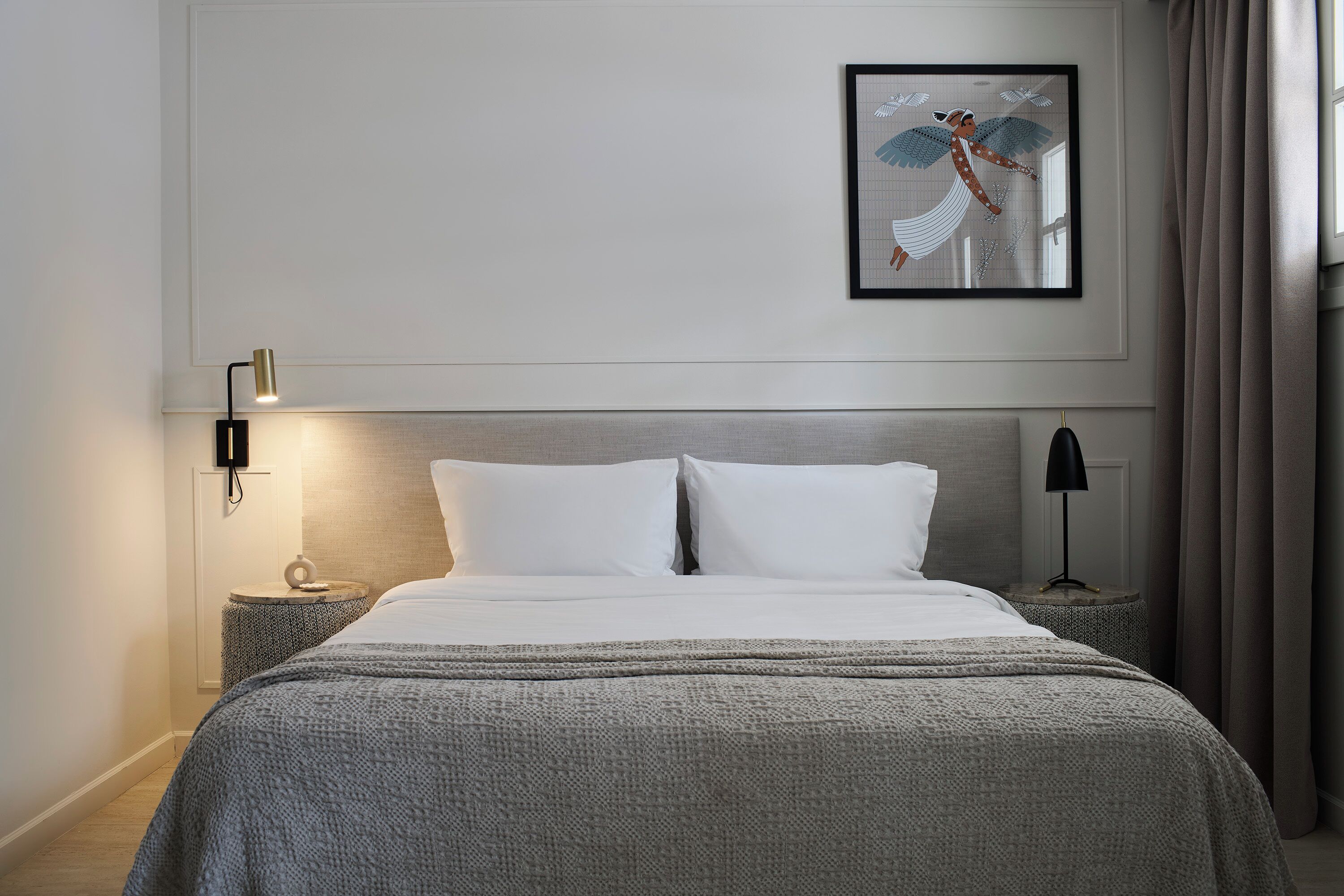 A neatly made bed with a grey blanket, two bedside tables with lamps, a framed artwork on the wall, and a window with curtains.