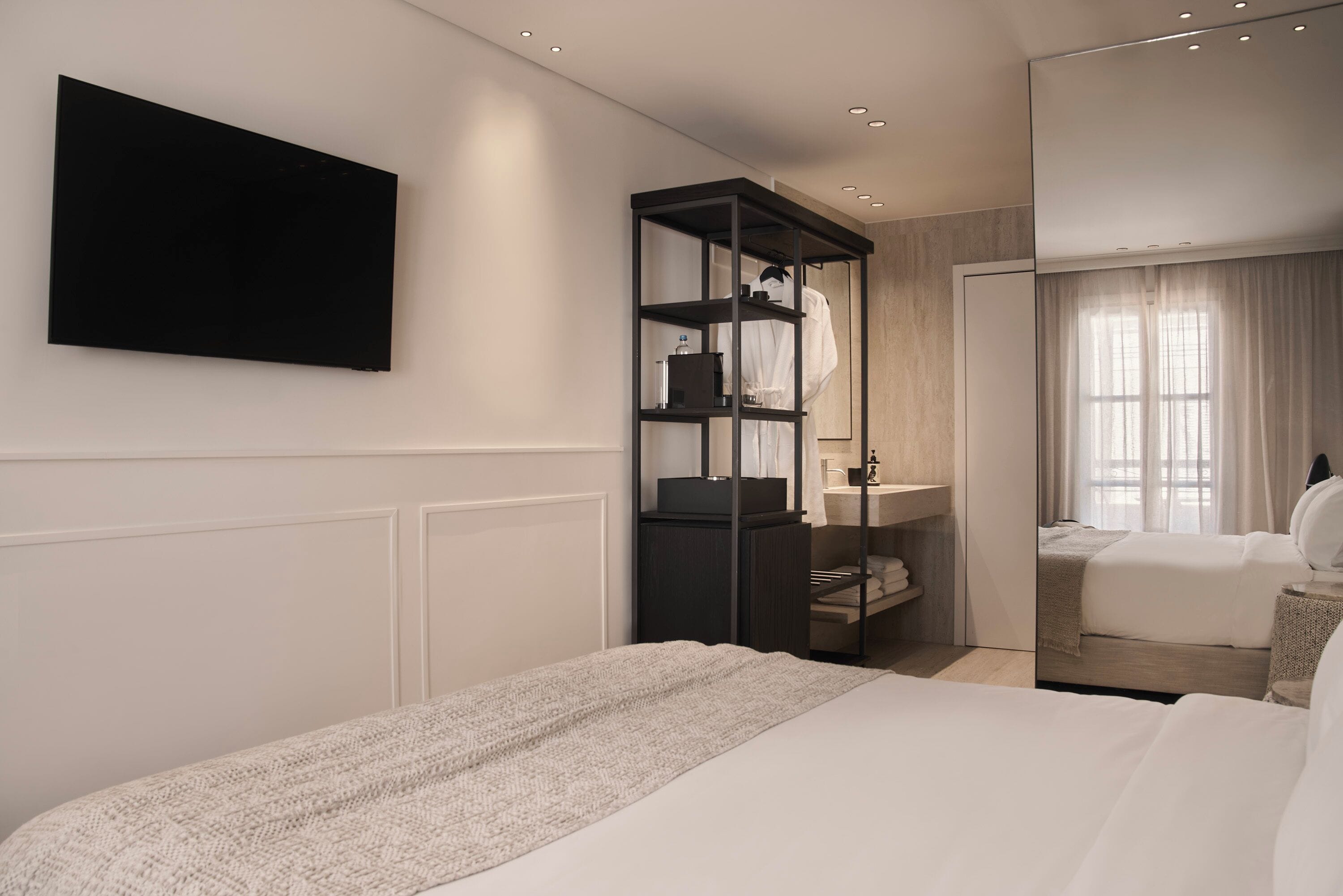 A modern hotel room with a large bed, a built-in TV, a wardrobe with shelves, and a bathroom with a mirror and curtains.