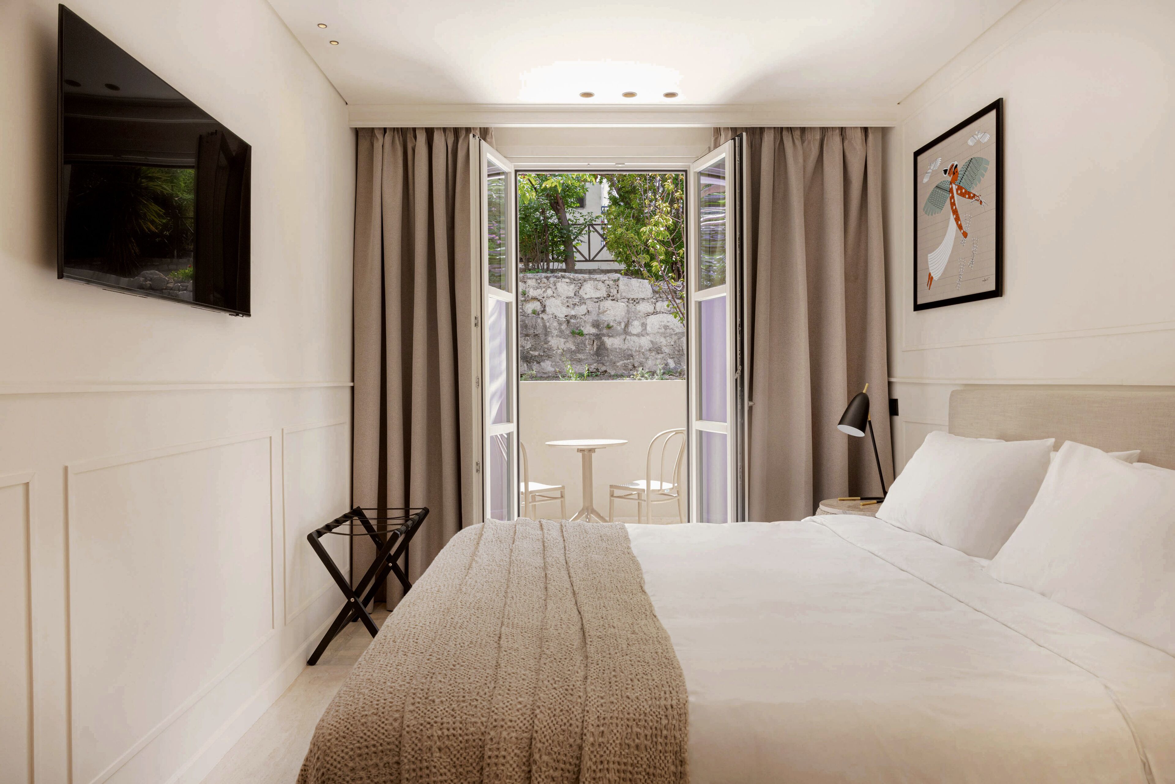 Habitación de hotel con cama, televisor montado en la pared, una mesita y sillas, y vista a un espacio exterior por una puerta abierta.