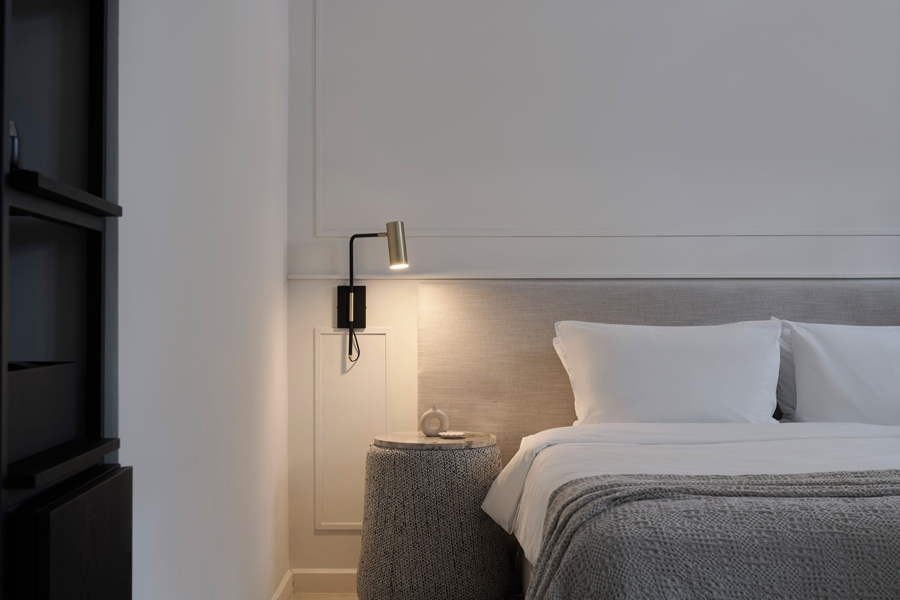 Una cama bien hecha con ropa blanca, una mesita redonda con lámpara y un estante fijado a la pared.