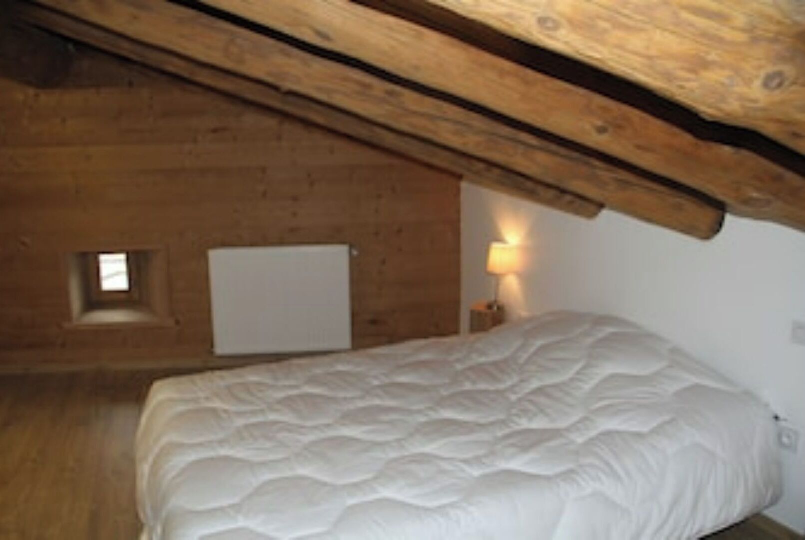 Quarto com teto inclinado, uma cama, mesa de cabeceira com abajur, pequena janela e radiador.