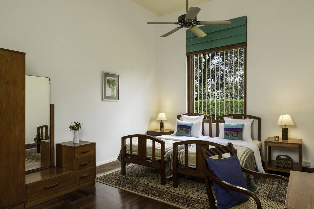 Un dormitorio con cama, dos mesitas de noche, una mesita auxiliar, una silla, cómoda, espejo, ventilador de techo y ventana con vistas a la vegetación.