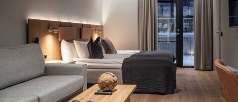 Una habitación de hotel moderna con sofá, cama con cabecera de madera, mesa de centro de madera con objetos decorativos, y vistas a través de una puerta de cristal.
