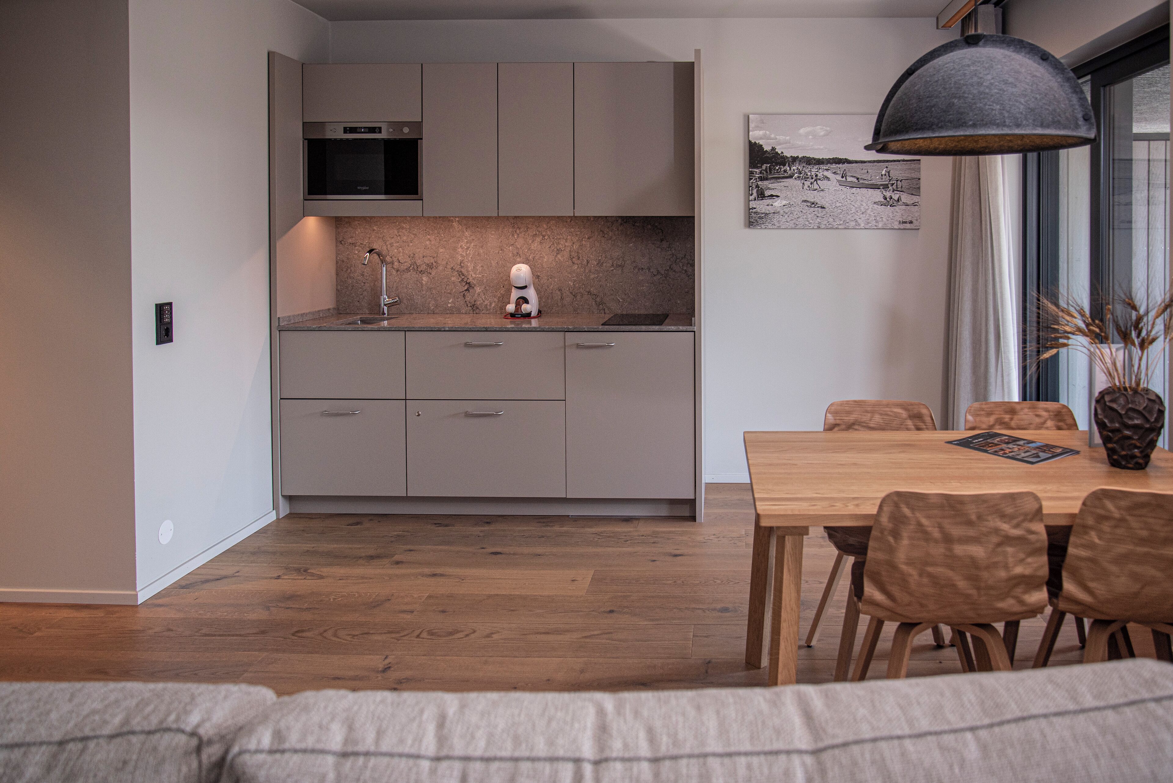 A modern kitchen with grey cabinets, a built-in oven, and a sink. There's a dining table with chairs and a pendant light above it.