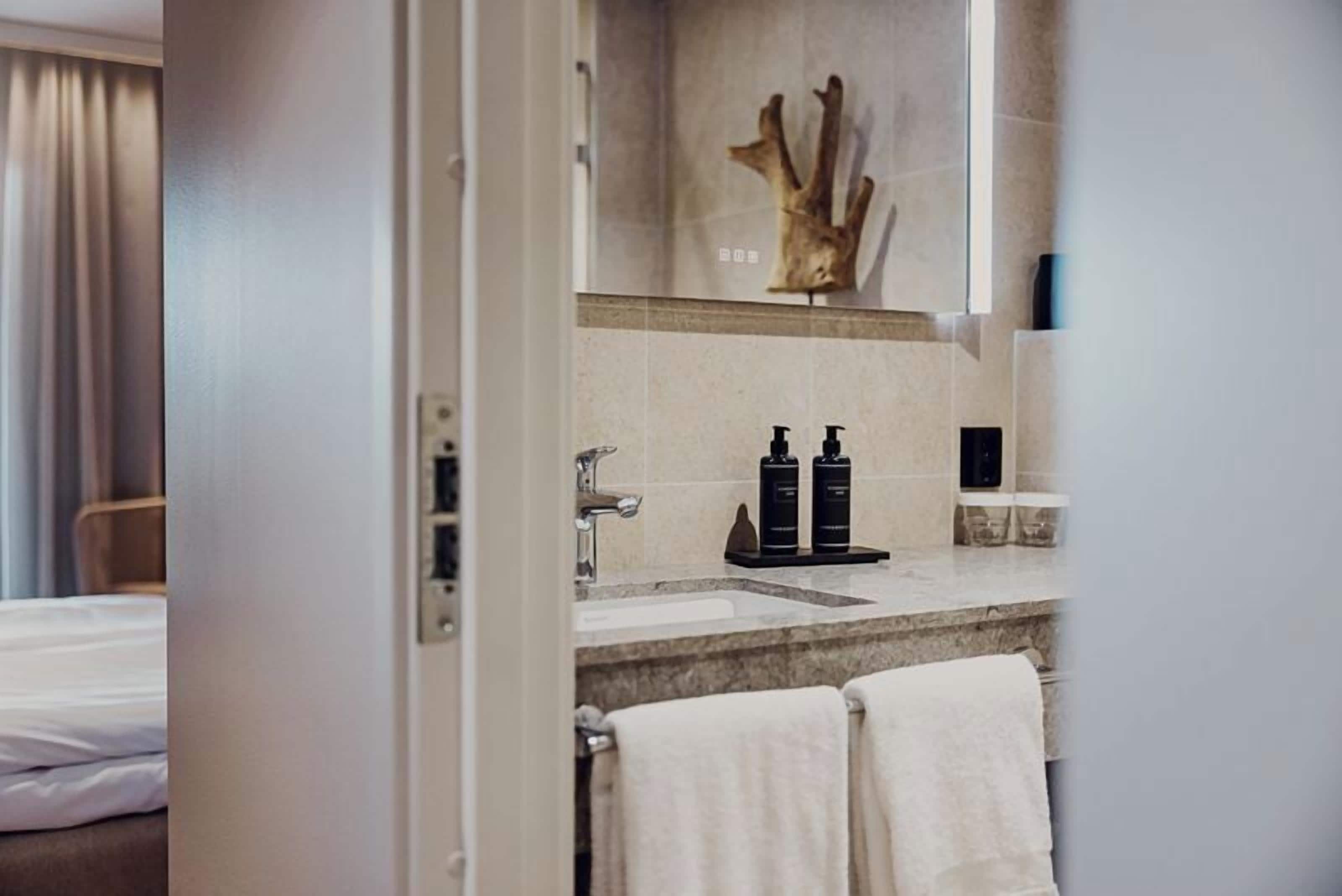 Baño de hotel con lavamanos, dos artículos de tocador negros, un perchero con dos toallas y un adorno de madera sobre el espejo.