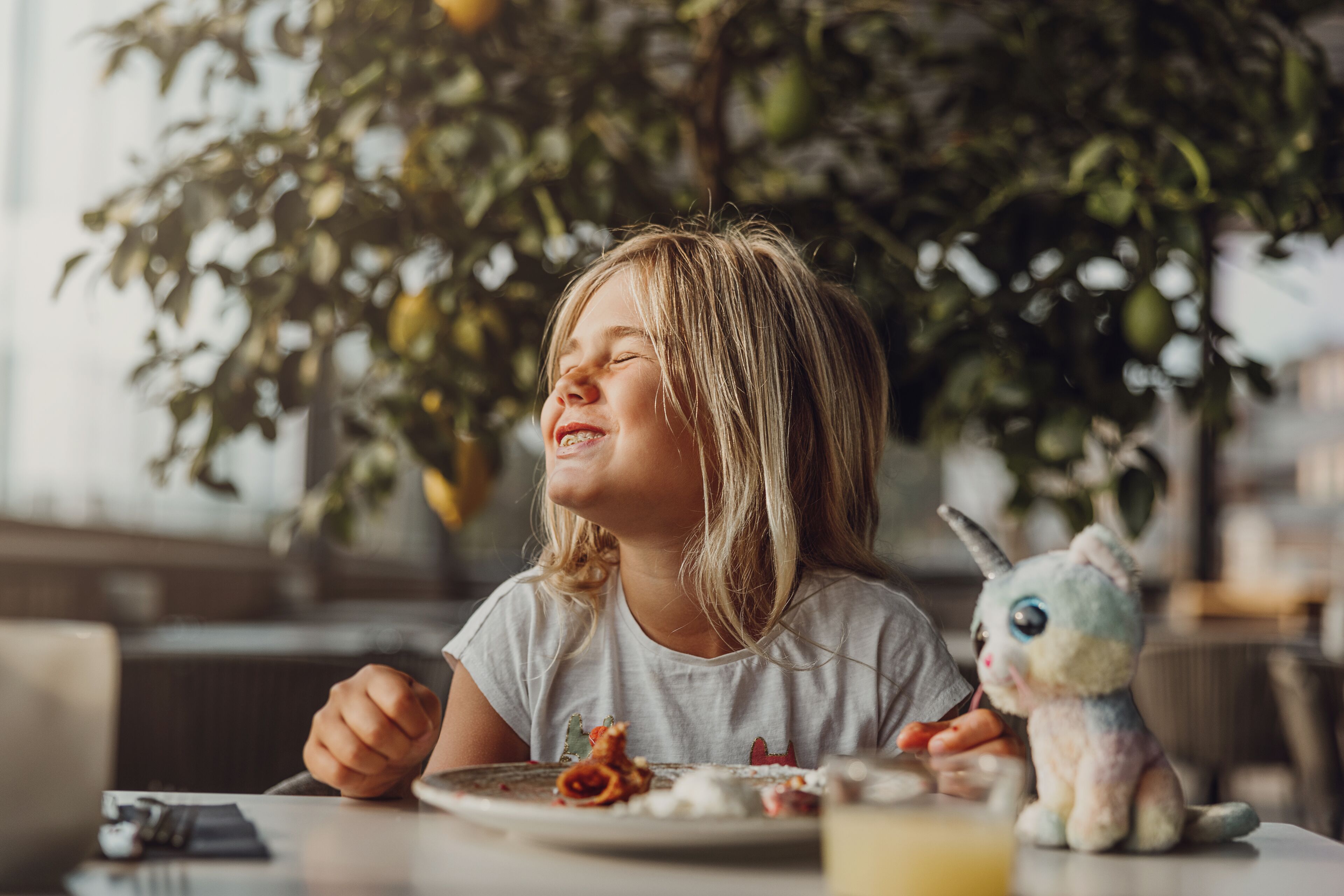 Een kind zittend aan een tafel met een bord eten, een knuffel en een glas sap.
