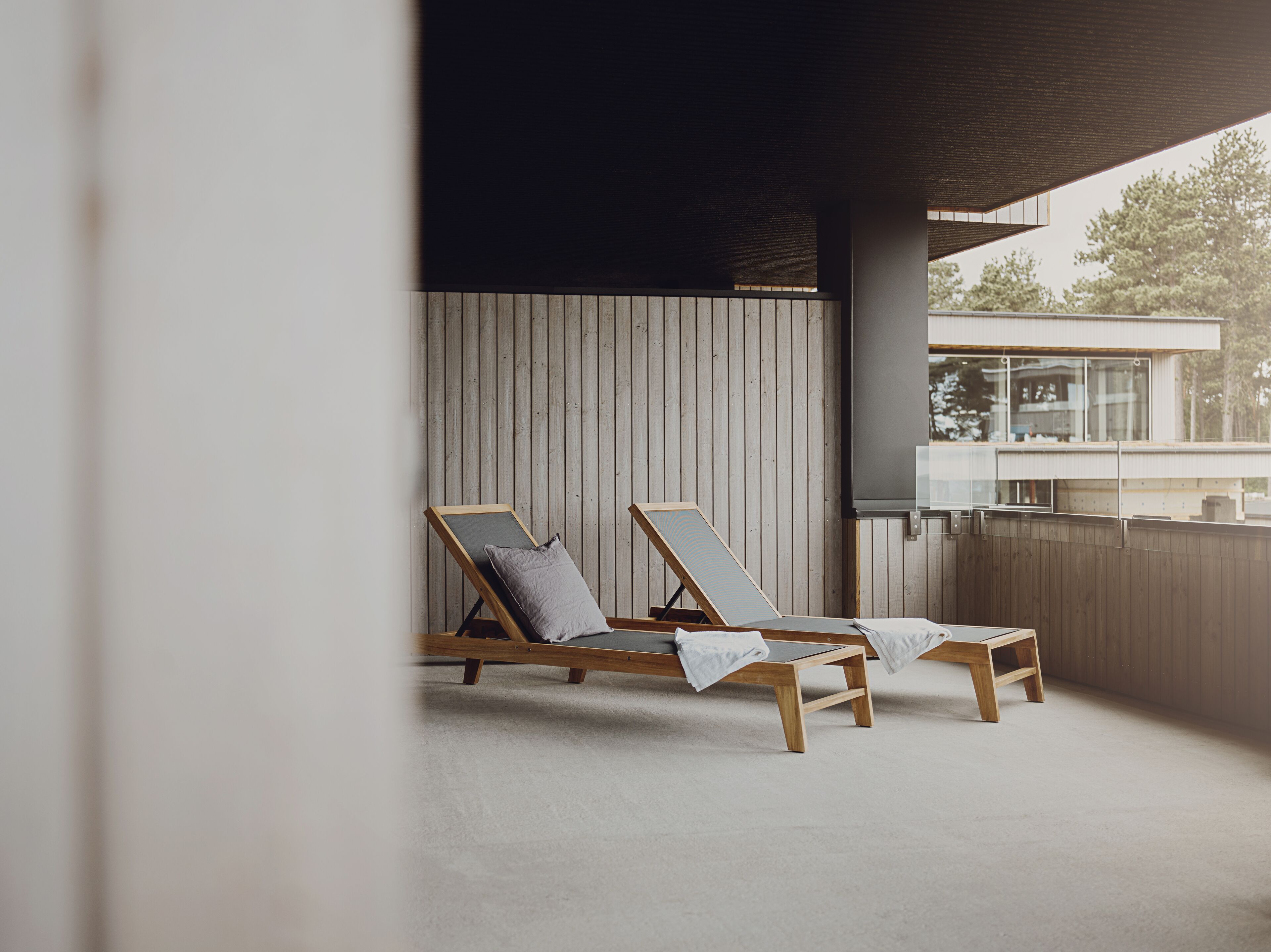 A modern outdoor lounge area with two wooden lounge chairs, a concrete floor, and a wooden wall.