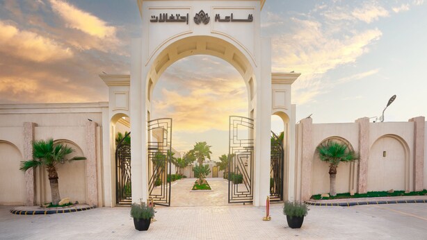 Une entrée ornée d’un arc et de motifs calligraphiques arabes, entourée de palmiers et bordée d’un chemin pavé.
