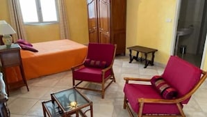 A room with a bed, two red chairs, a small table, a cabinet, and a window.