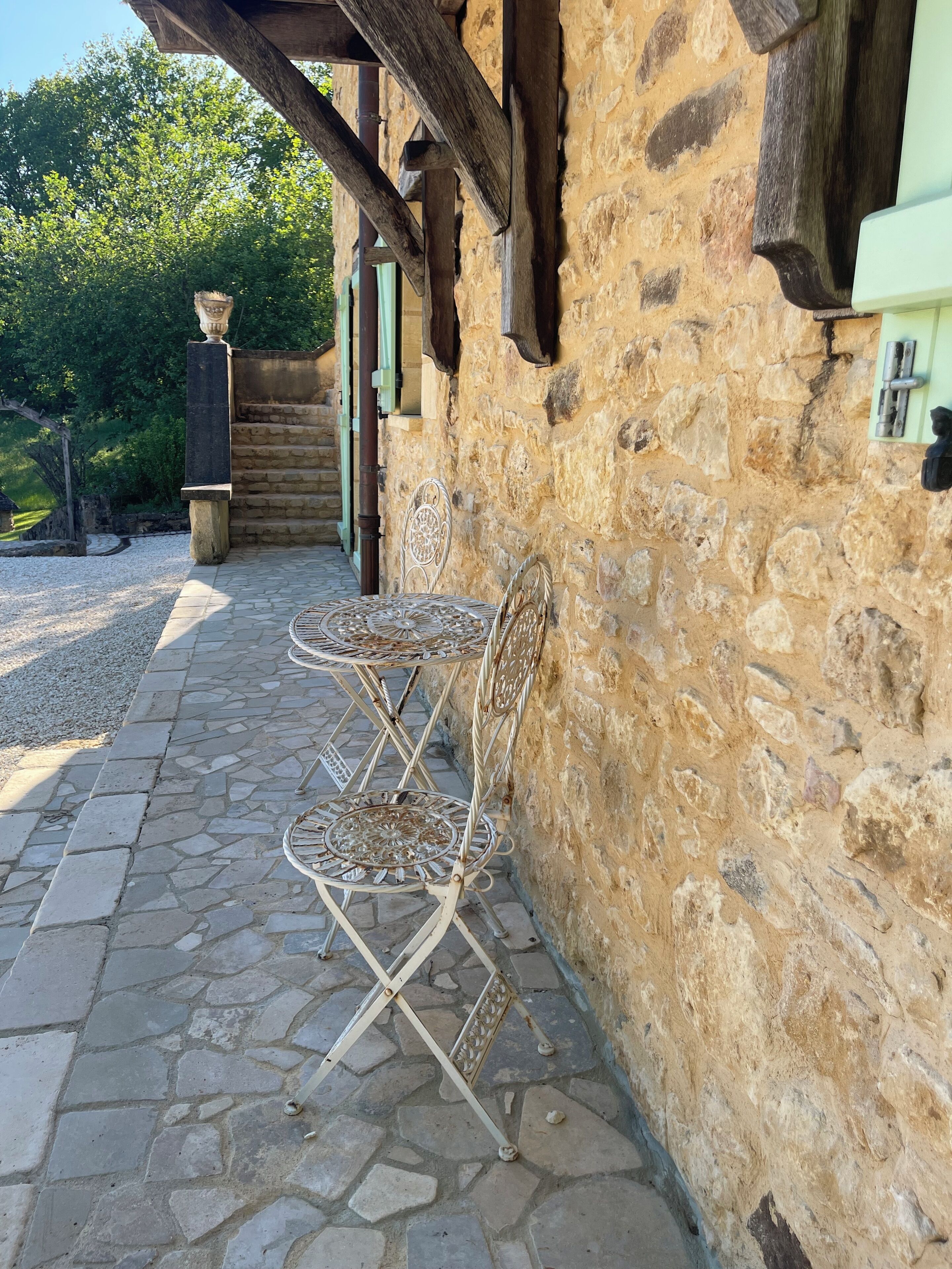Een stenen terras met metalen stoelen en een tafel, een stenen muur en een houten constructie op de achtergrond.
