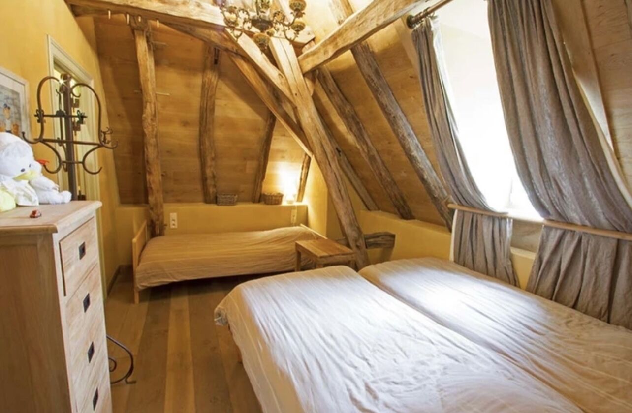 A rustic attic bedroom with exposed wooden beams, a chandelier, and a large window with curtains.