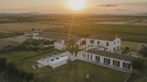 Una casa grande con piscina y varias habitaciones, rodeada de campos y un atardecer al fondo.