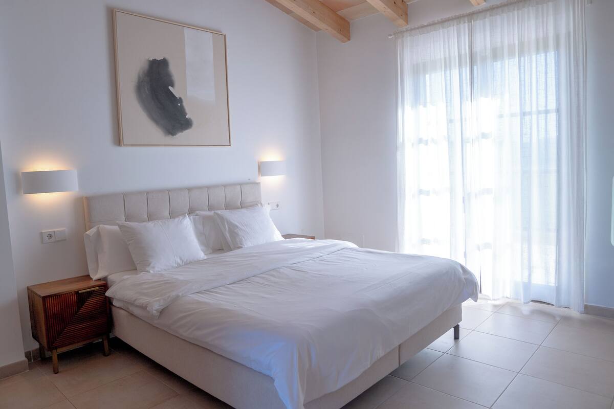 A neatly made bed in a room with a large window, a framed abstract painting, and two wall-mounted lamps.