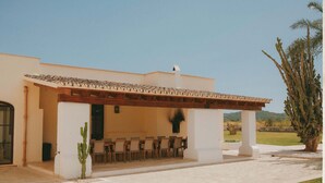 Un área de estar al aire libre techada con sillas y una mesa, una planta de cactus y un árbol grande al fondo.