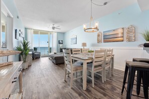 Une salle à manger spacieuse, avec une table et des chaises en bois, un ventilateur de plafond et une vue sur l’océan grâce à de grandes fenêtres.