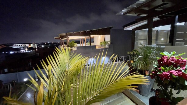 A rooftop terrace with a view of the city at night, featuring a modern building with a glass railing and potted plants.
