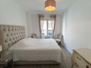 Une chambre à coucher avec une tête de lit capitonnée, un grand lit, une table de chevet, une chaise, une commode et une fenêtre avec des rideaux.
