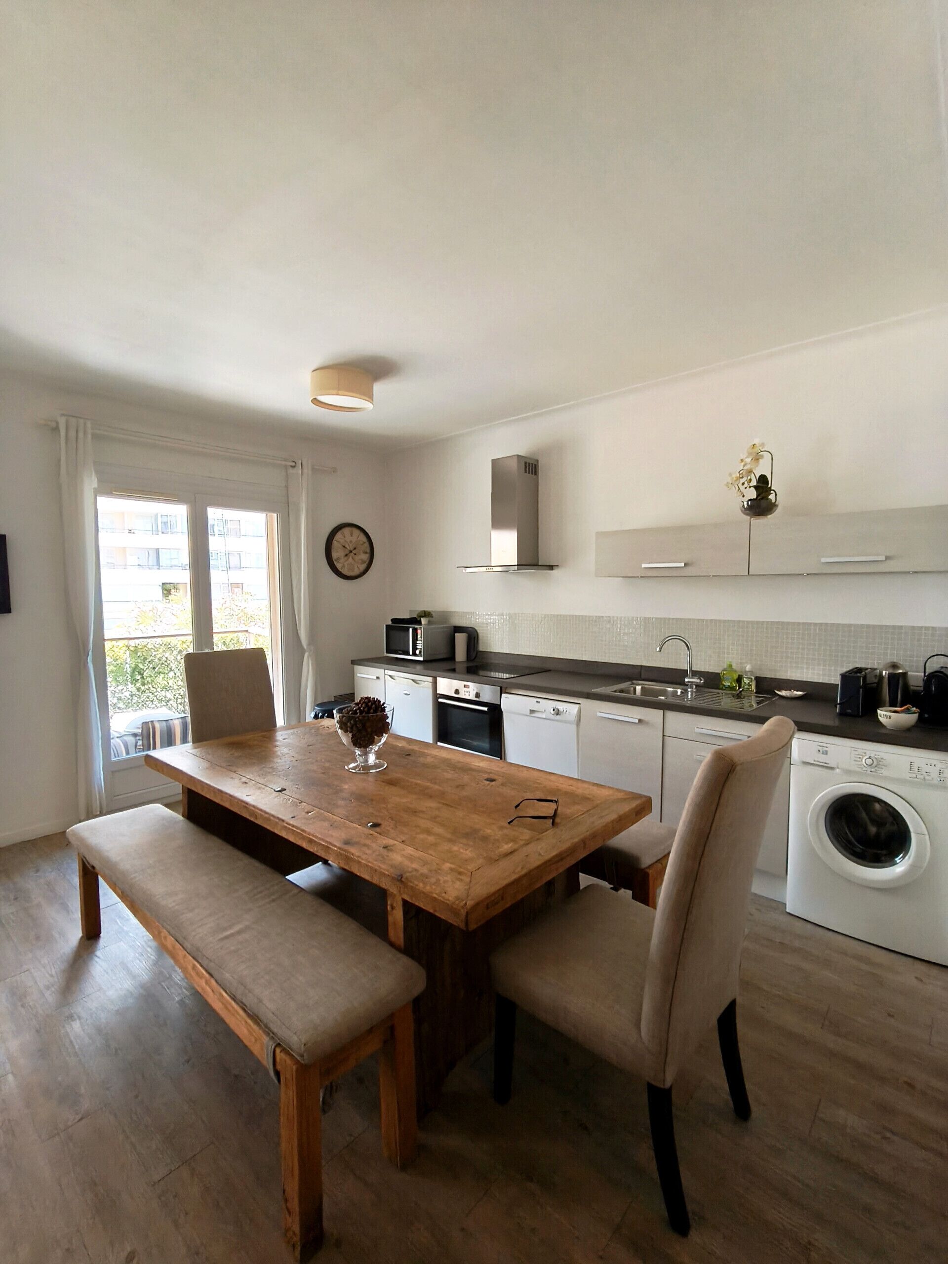 Een moderne keuken met een houten eettafel, stoelen, een wasmachine en uitzicht op buiten.