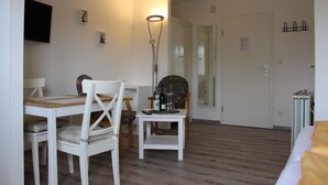 A room with a wooden floor, white walls, a dining table with chairs, a standing lamp, and a bed with a yellow blanket.