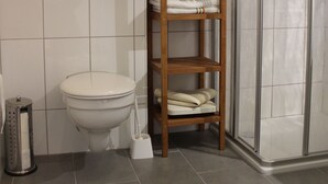 A bathroom with a toilet, a wooden shelf with towels, a shower, and a tiled wall with a light fixture.