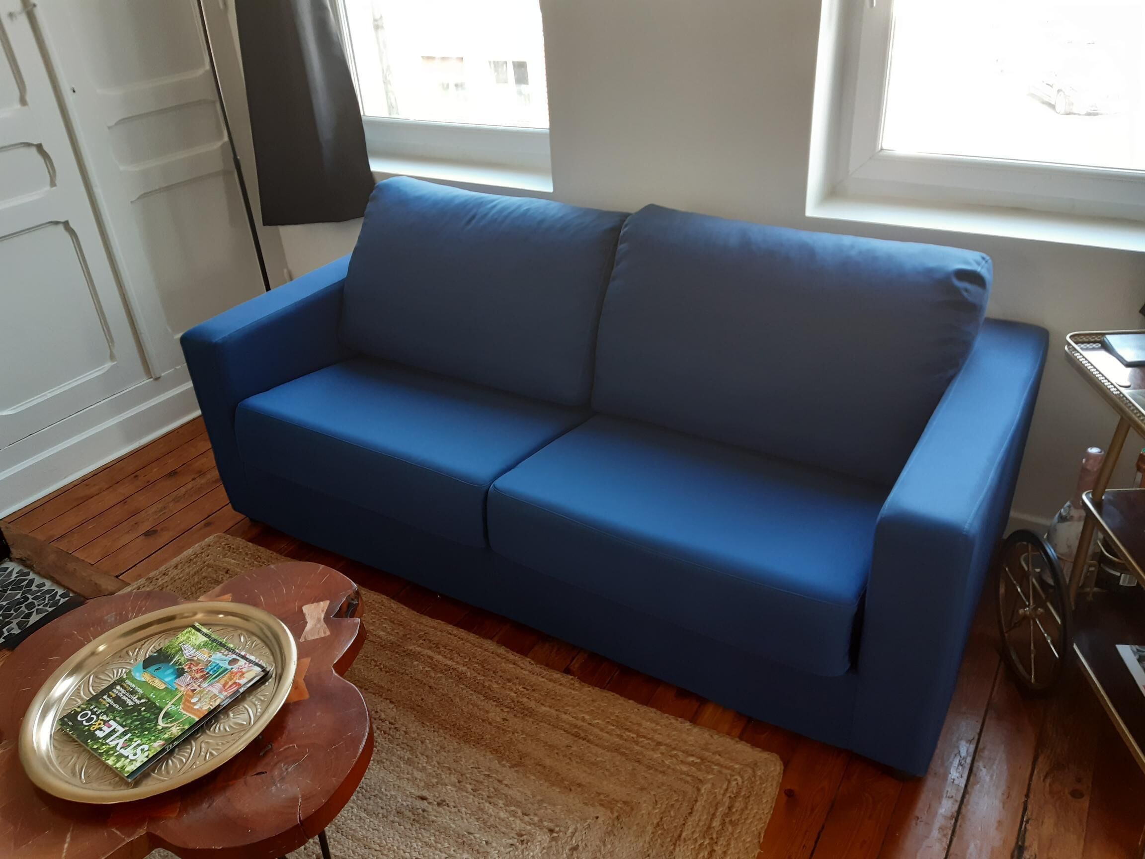 En blå soffa i ett rum med trägolv, ett litet runt bord med ett tidningsnummer på och ett fönster med gardiner.