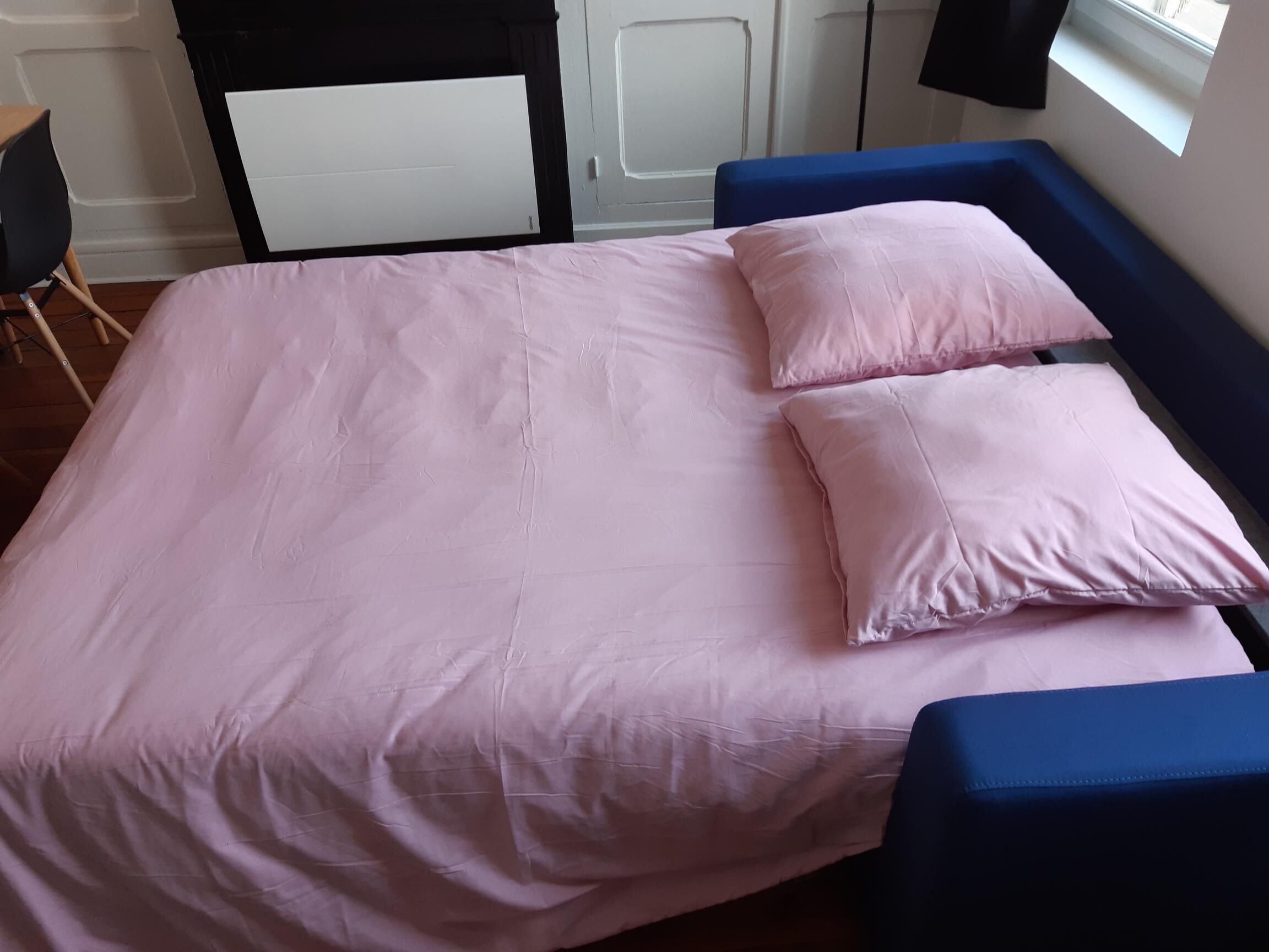 Ein Bett mit einer rosa Bettdecke und Kissen, einem blauen Kopfteil und einem Fenster mit Tageslicht.