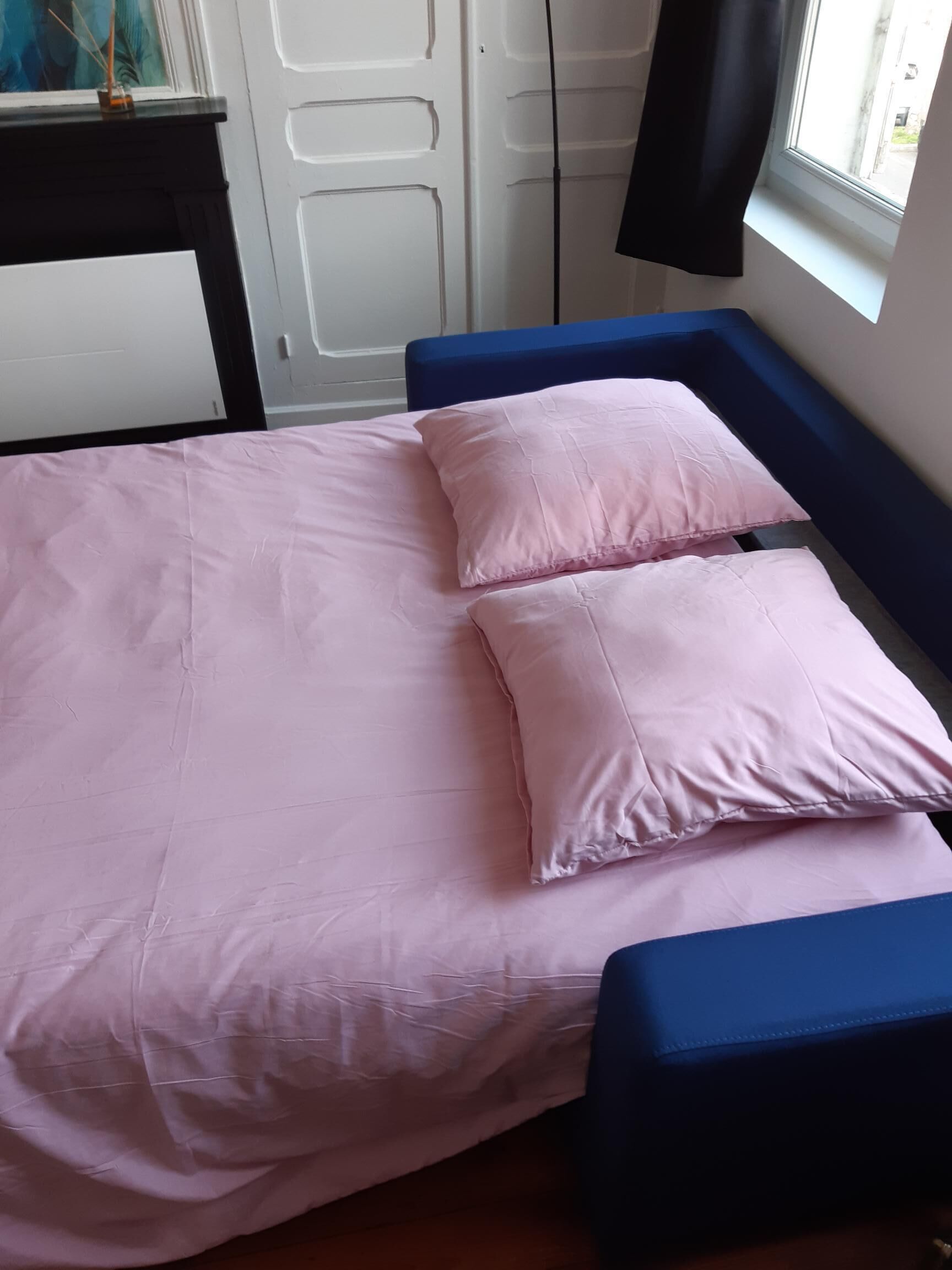 Un lit avec une couvre-lit et des oreillers roses, une tête de lit bleue et une fenêtre donnant sur l’extérieur.