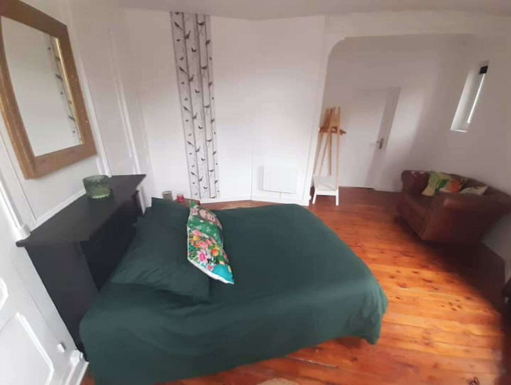 A bedroom with a green bedspread, a dresser, a mirror, a window with a curtain, and a wooden floor.