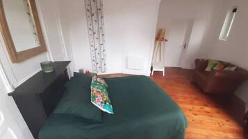 A bedroom with a green bedspread, a dresser, a mirror, a window with a curtain, and a wooden floor.