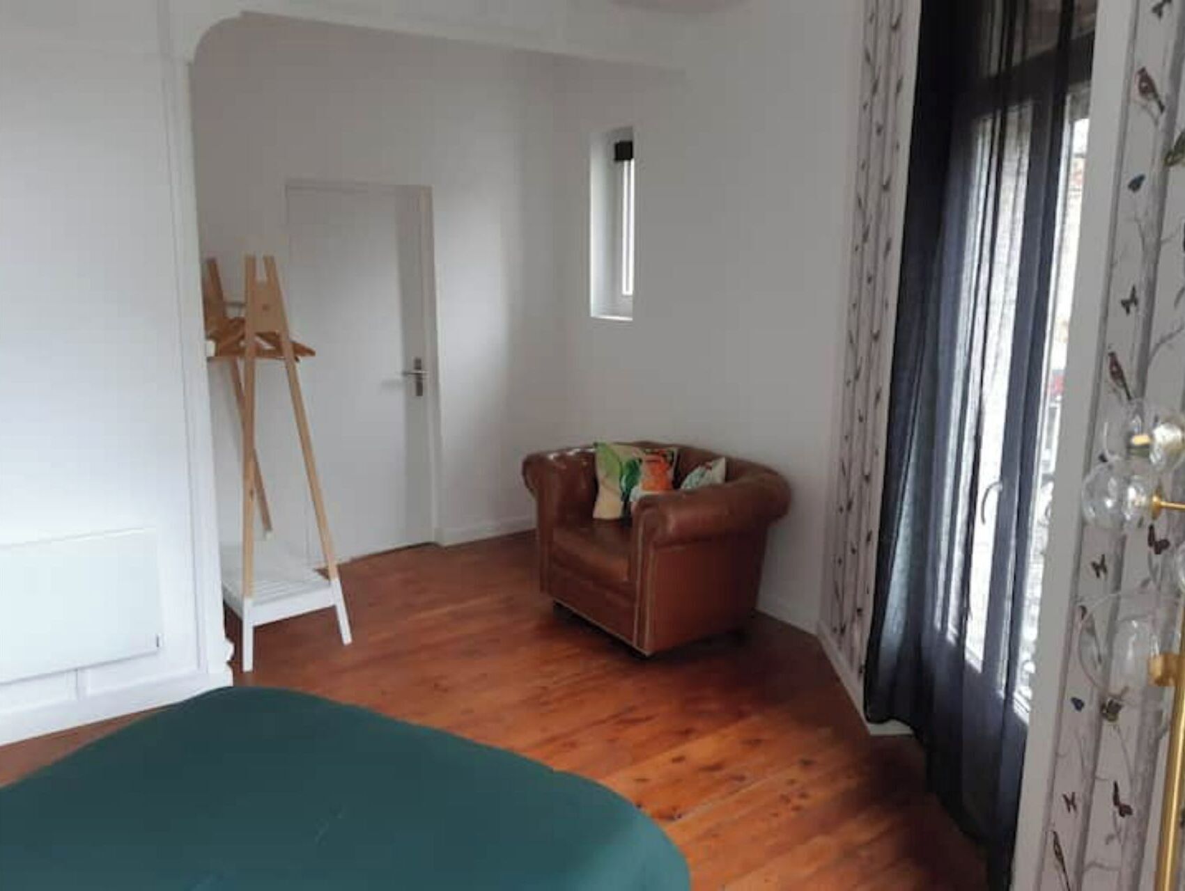 A room with a wooden floor, a brown leather sofa, a green ottoman, a white door, and a window with curtains.