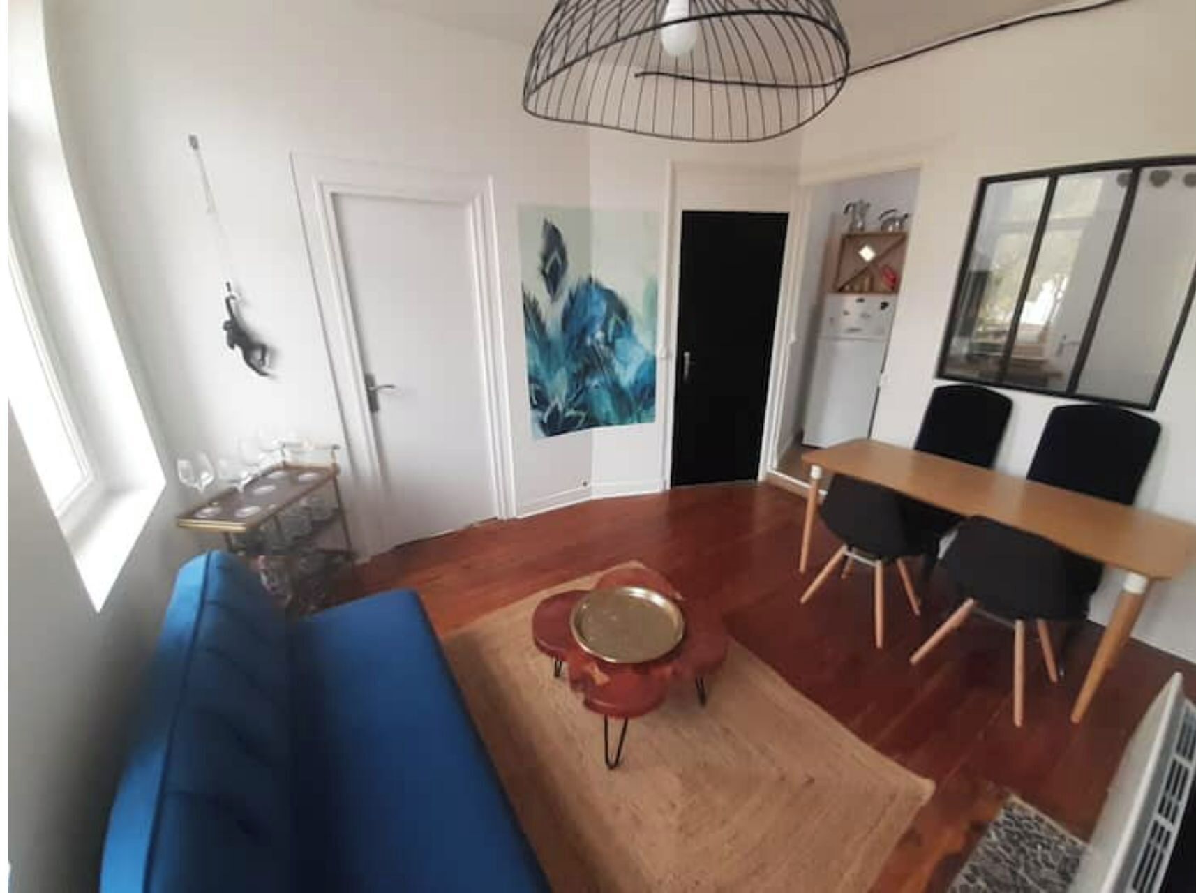 A modern living room with a blue sofa, a dining table with chairs, a kitchen area, and a large window.