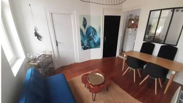 A modern living room with a blue sofa, a dining table with chairs, a kitchen area, and a large window.
