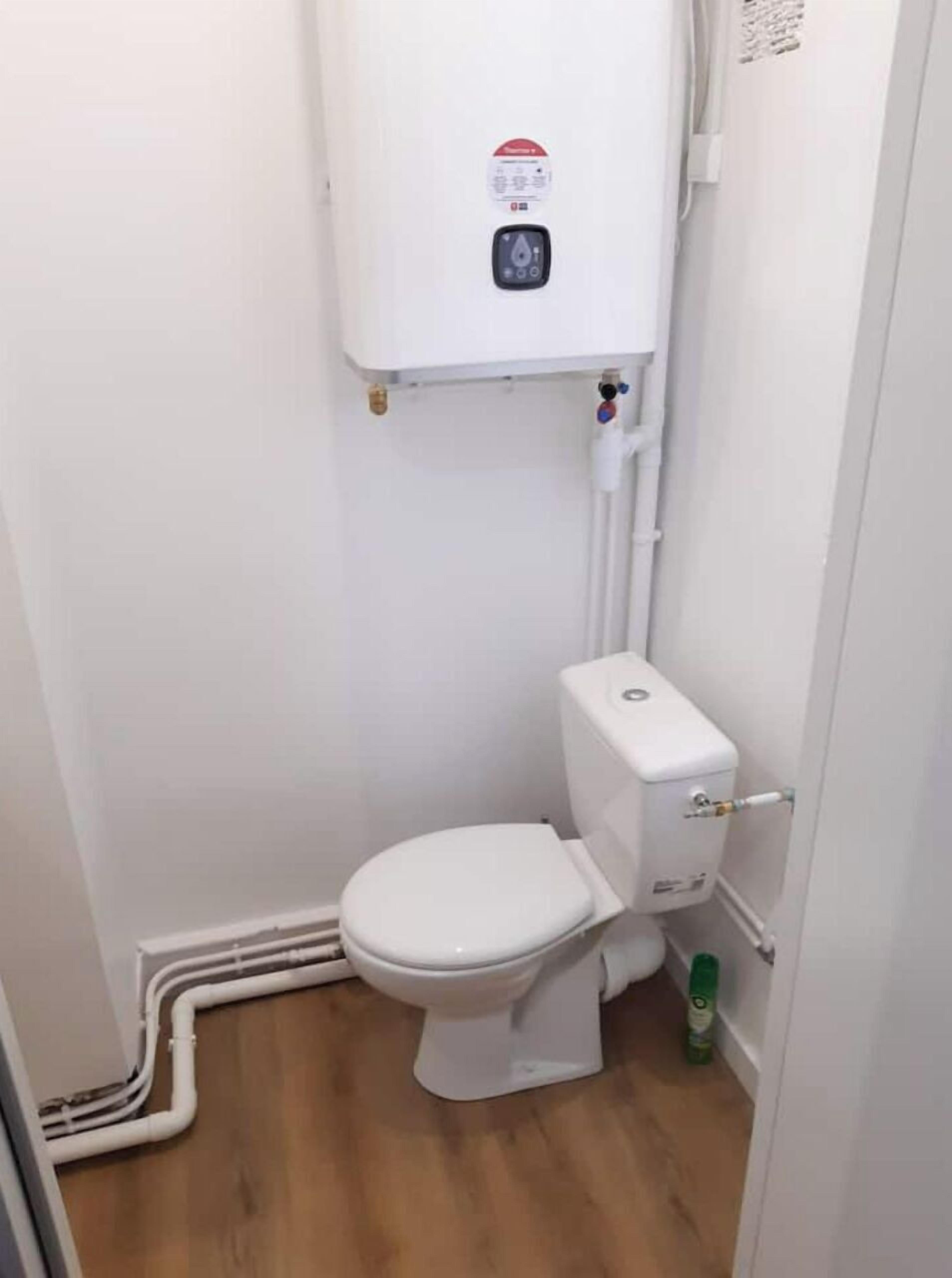 Une petite salle de bain blanche, avec des toilettes, une cuvette blanche et des tuyaux apparents.