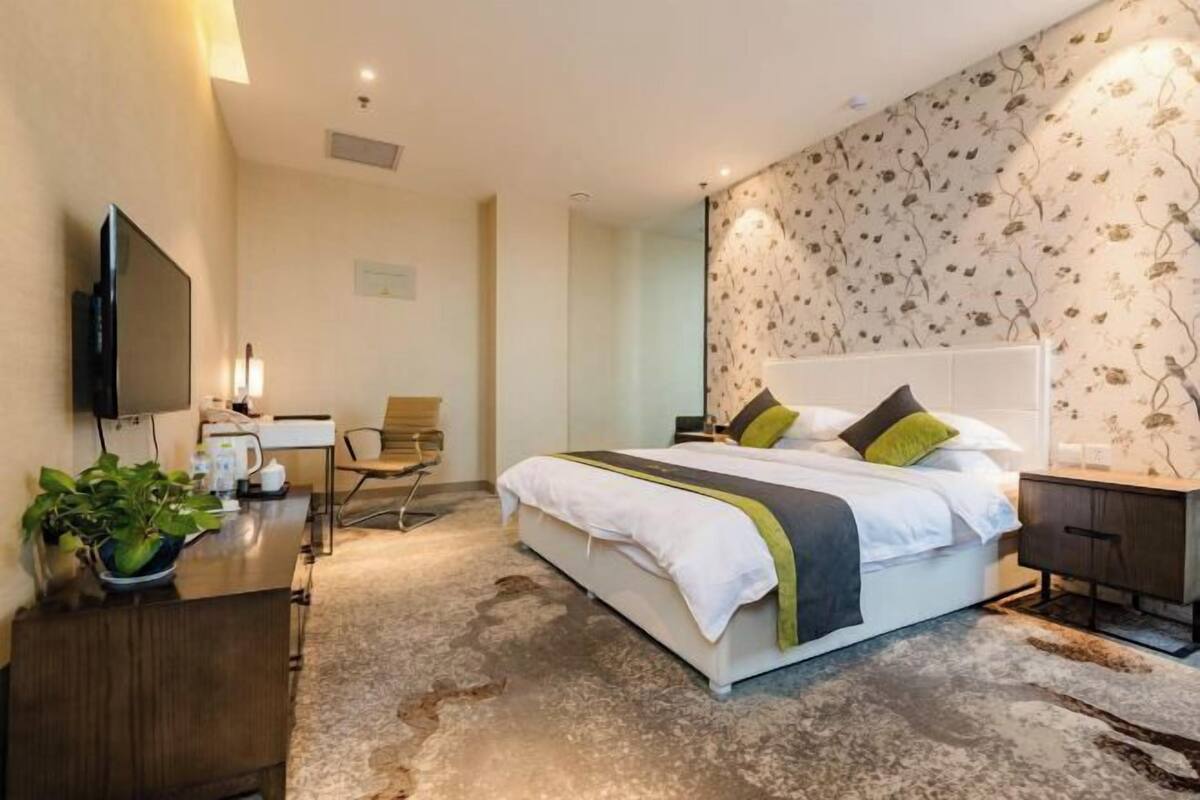A hotel room with a bed, a desk with a flat-screen TV, a chair, and a potted plant.