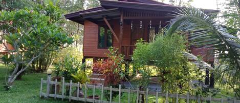A wooden cabin with a sloped roof, surrounded by lush greenery and a wooden fence.