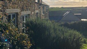 A stone building with multiple windows and a chimney, surrounded by greenery and a view of the landscape. - Cosy cottage, peaceful setting 5 mins to beautiful sandy beach & fabulous walks! (Mawgan Porth, Cornwall)