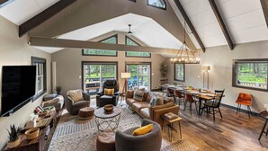 A spacious living room with a high ceiling, large windows, and a dining area with modern furniture. - PBJ Chalet – Modern Pocono Stay w/ Game Room, EV Charger & Fireplaces (Pocono Lake)