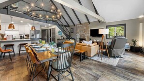 A spacious living area with a dining table, chairs, a fireplace, and a large window. - PBJ Chalet – Modern Pocono Stay w/ Game Room, EV Charger & Fireplaces (Pocono Lake)