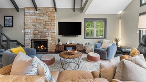 A cozy living room with a stone fireplace, comfortable seating, a flat-screen TV, and a large window. - PBJ Chalet – Modern Pocono Stay w/ Game Room, EV Charger & Fireplaces (Pocono Lake)