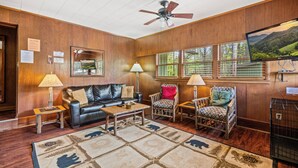 A living room with a leather sofa, two armchairs, a coffee table, a ceiling fan, a TV, and a window with a view of greenery.