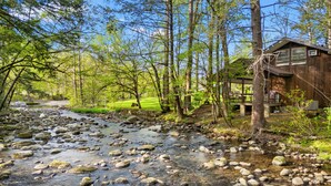 A cabin with a porch, a stream with rocks, and a forested area.