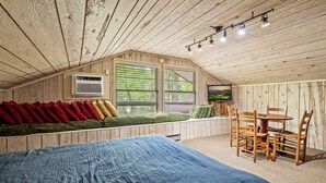 A cozy room with a sloped ceiling, a bed, a seating area with cushions, a dining table with chairs, and a window with blinds.