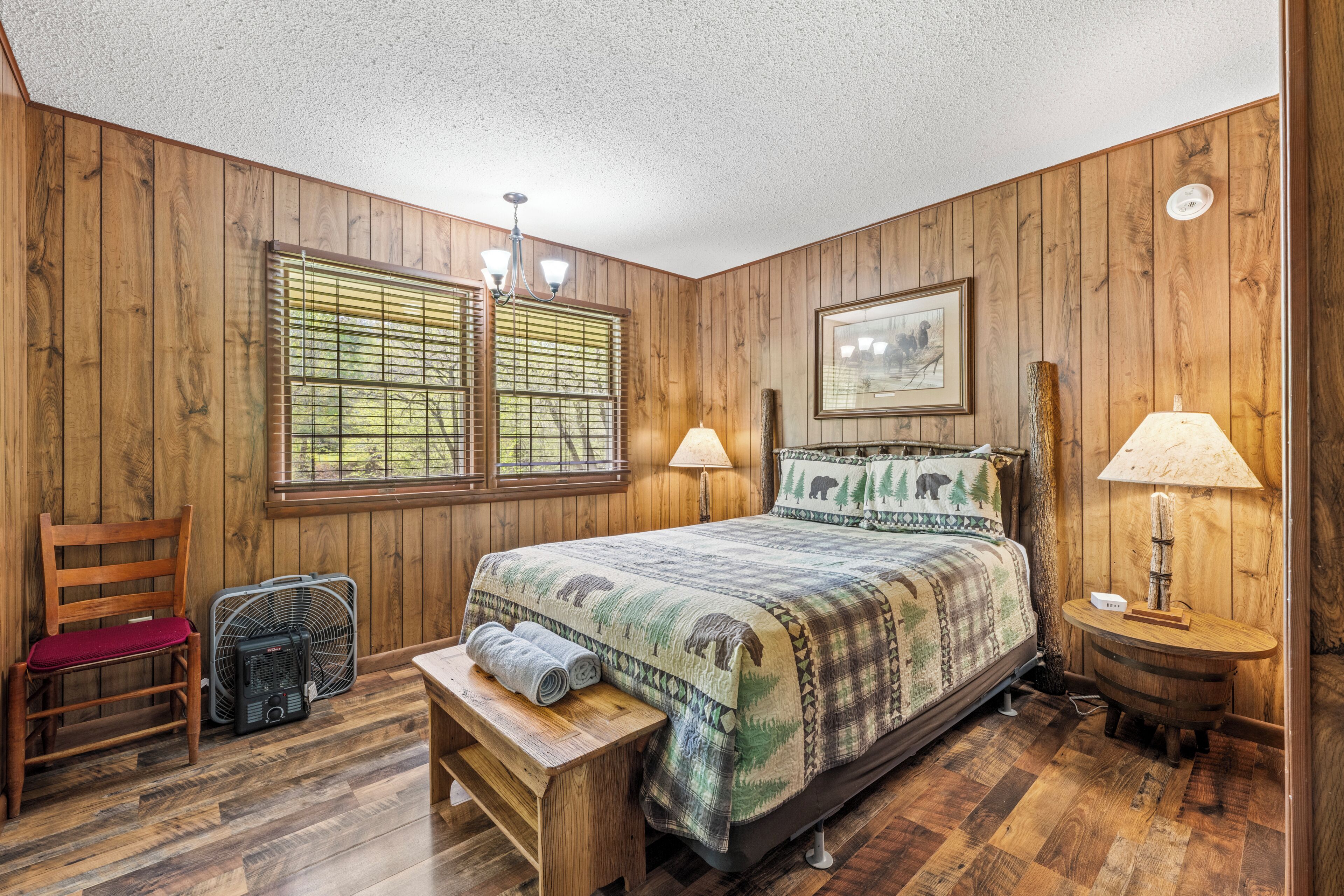 A wooden-paneled room with a bed, a chair, a bench, a fan, and a lamp.