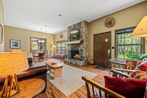 A cozy living room with a stone fireplace, wooden furniture, and large windows.