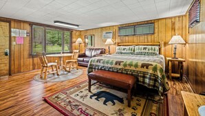 A wooden-paneled room with a bed, a wooden bench, a dining table with chairs, and a window with a view of trees.
