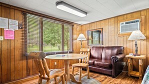 A wooden-paneled room with a round table, two chairs, a brown leather sofa, and a lamp on a wooden side table.