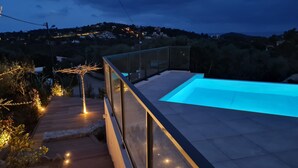 En moderne udendørs swimmingpool med glasgelænder, omgivet af en træterrasse og oplyst af lyskæder.