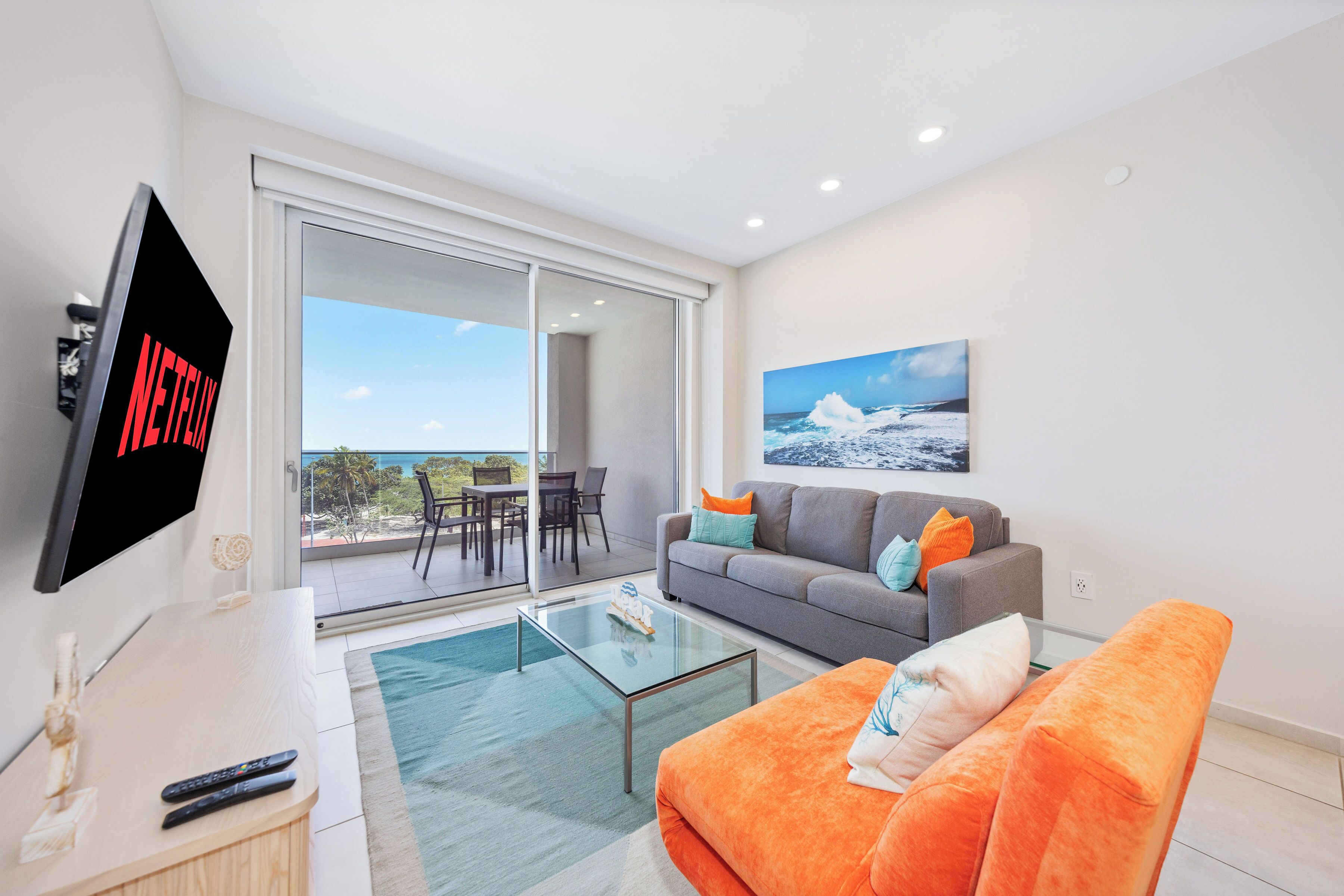 A modern living room with a flat-screen TV, a gray sofa, an orange couch, and a glass coffee table.